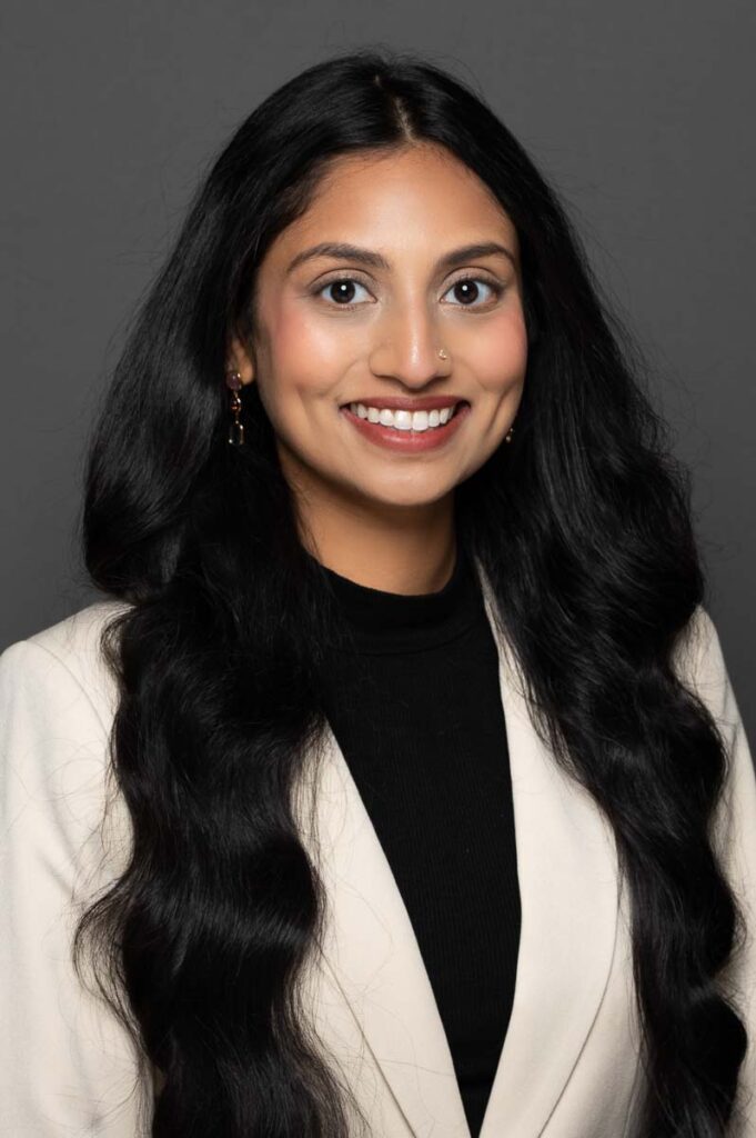 Headshot of Indian woman in white jacket on dark grey backdrop.