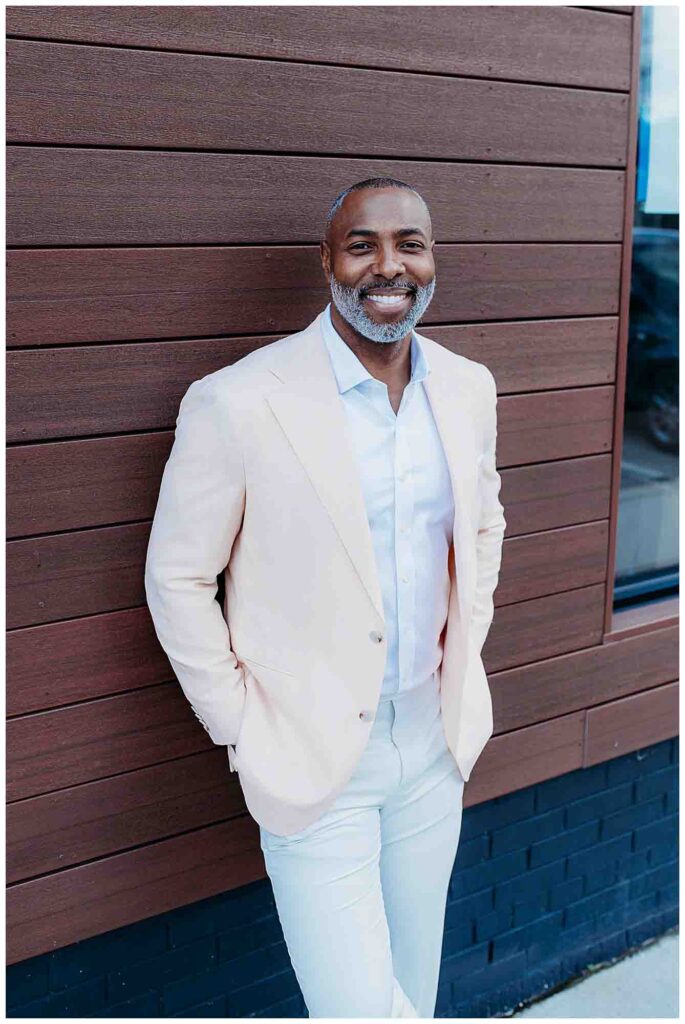 Man with pink sportcoat smiling at camera leaning against wood wall