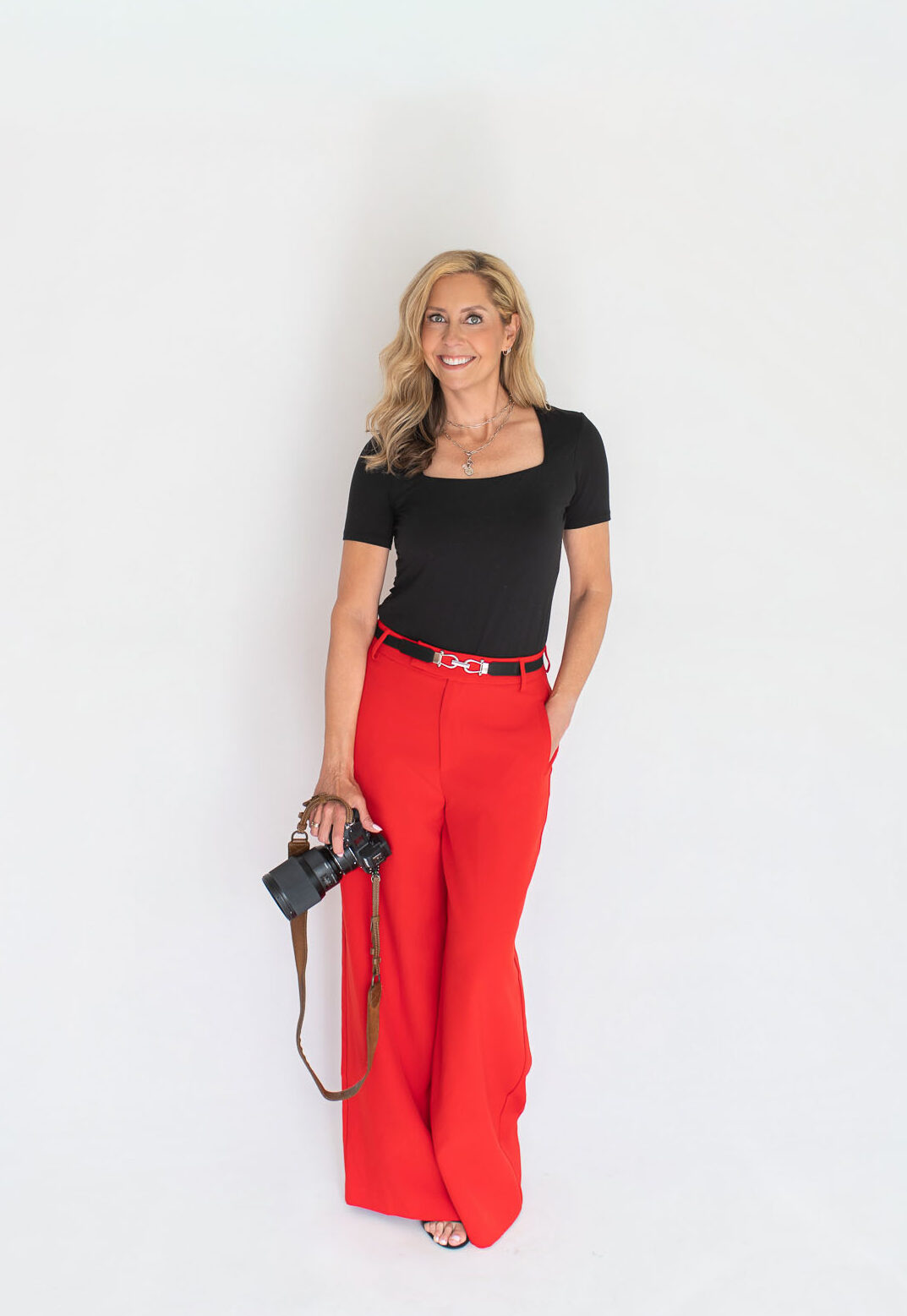Chicago Photographer in red pants and black top holding camera ready to take your headshot.